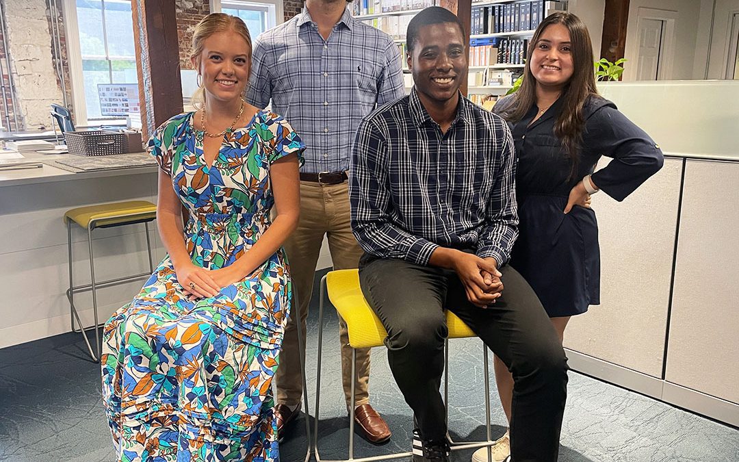 A photo of Garvin Design Group's four summer 2024 interns sitting and standing in front of a wall of bookshelves