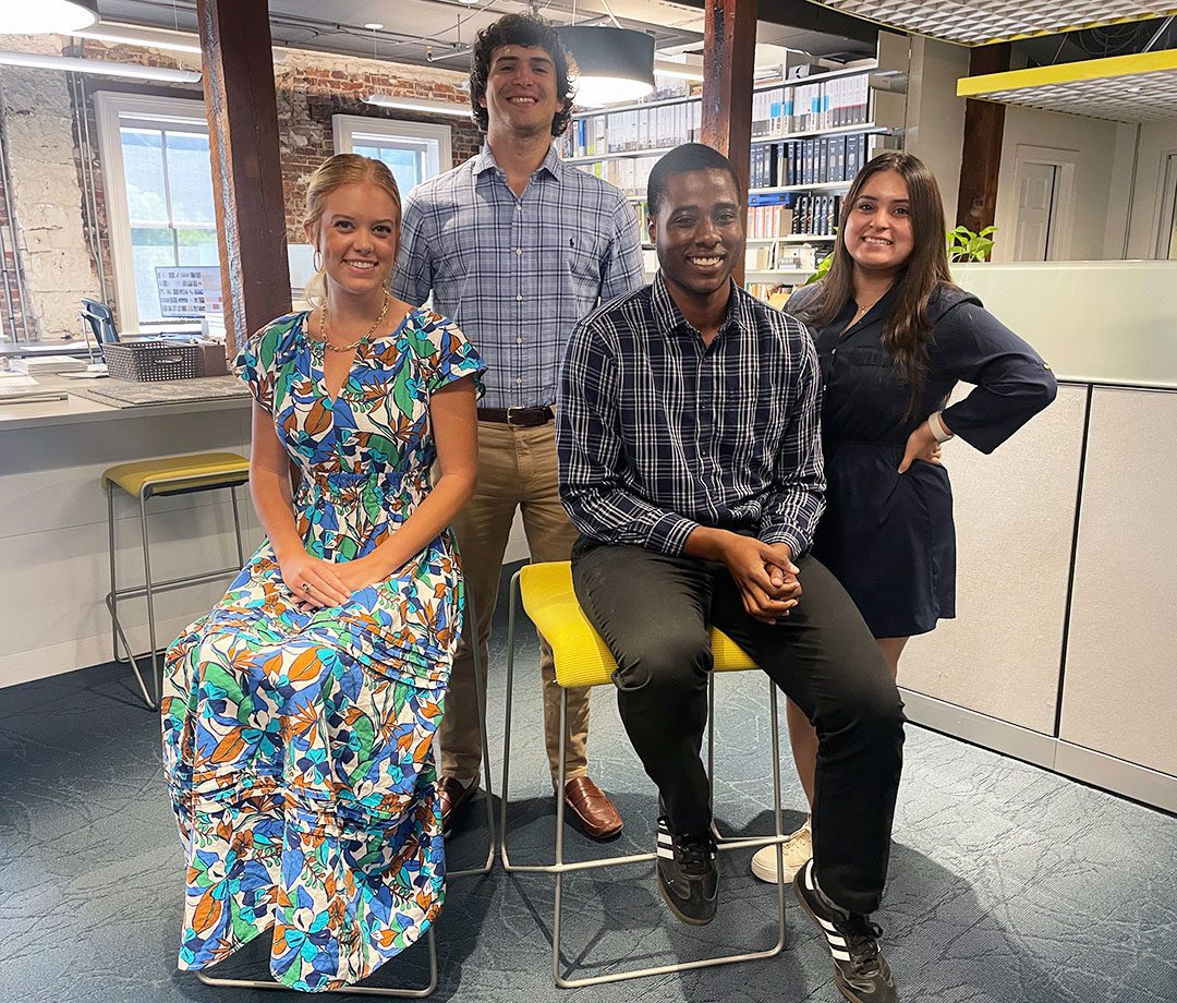 A photo of Garvin Design Group's four summer 2024 interns sitting and standing in front of a wall of bookshelves