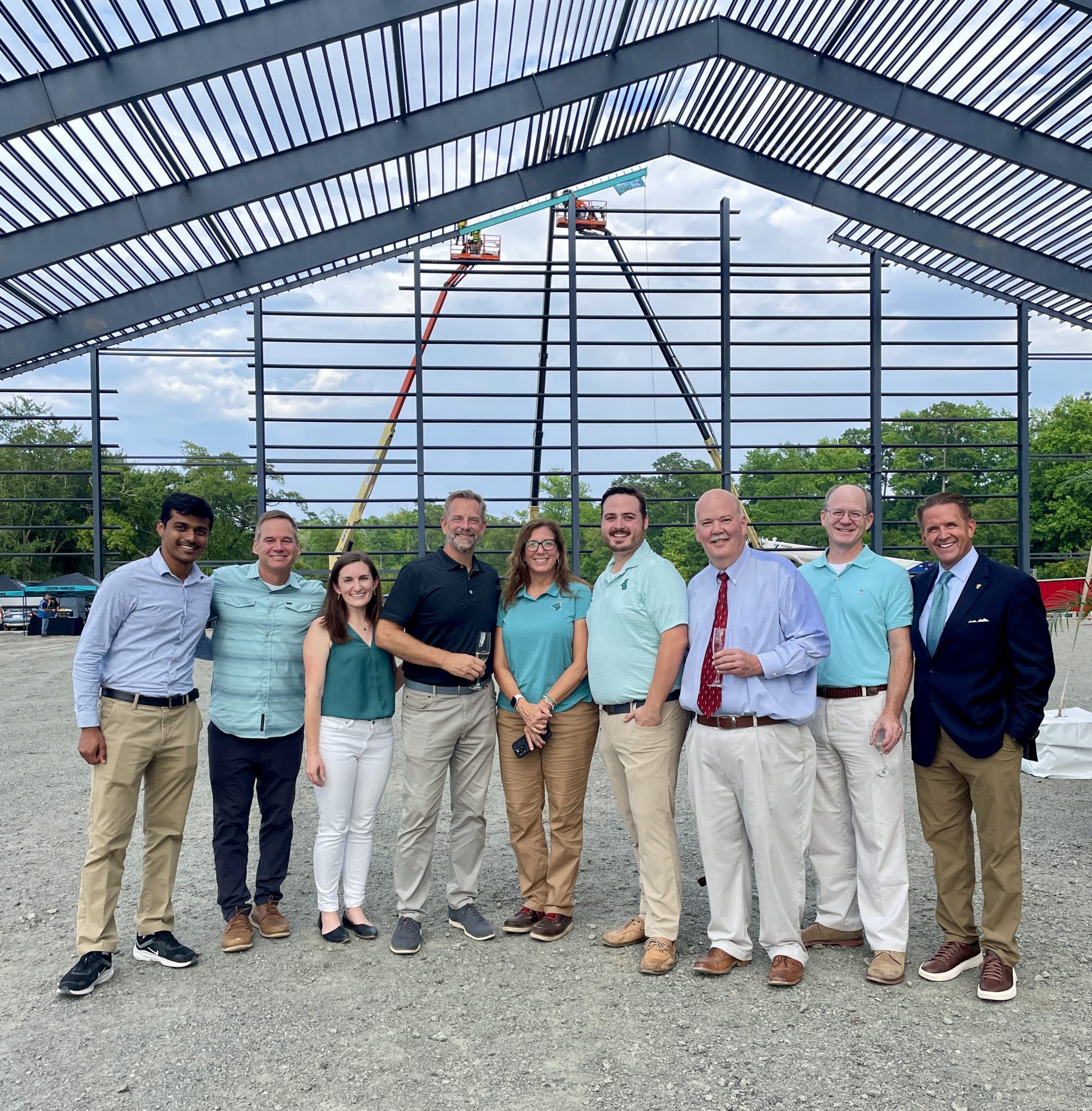 A photo of the project team at the topping out ceremony for Coastal Carolina University's Indoor Practice Facility. The team is a group of people standing inside a metal structure while a beam is lifted and set into place in the background.