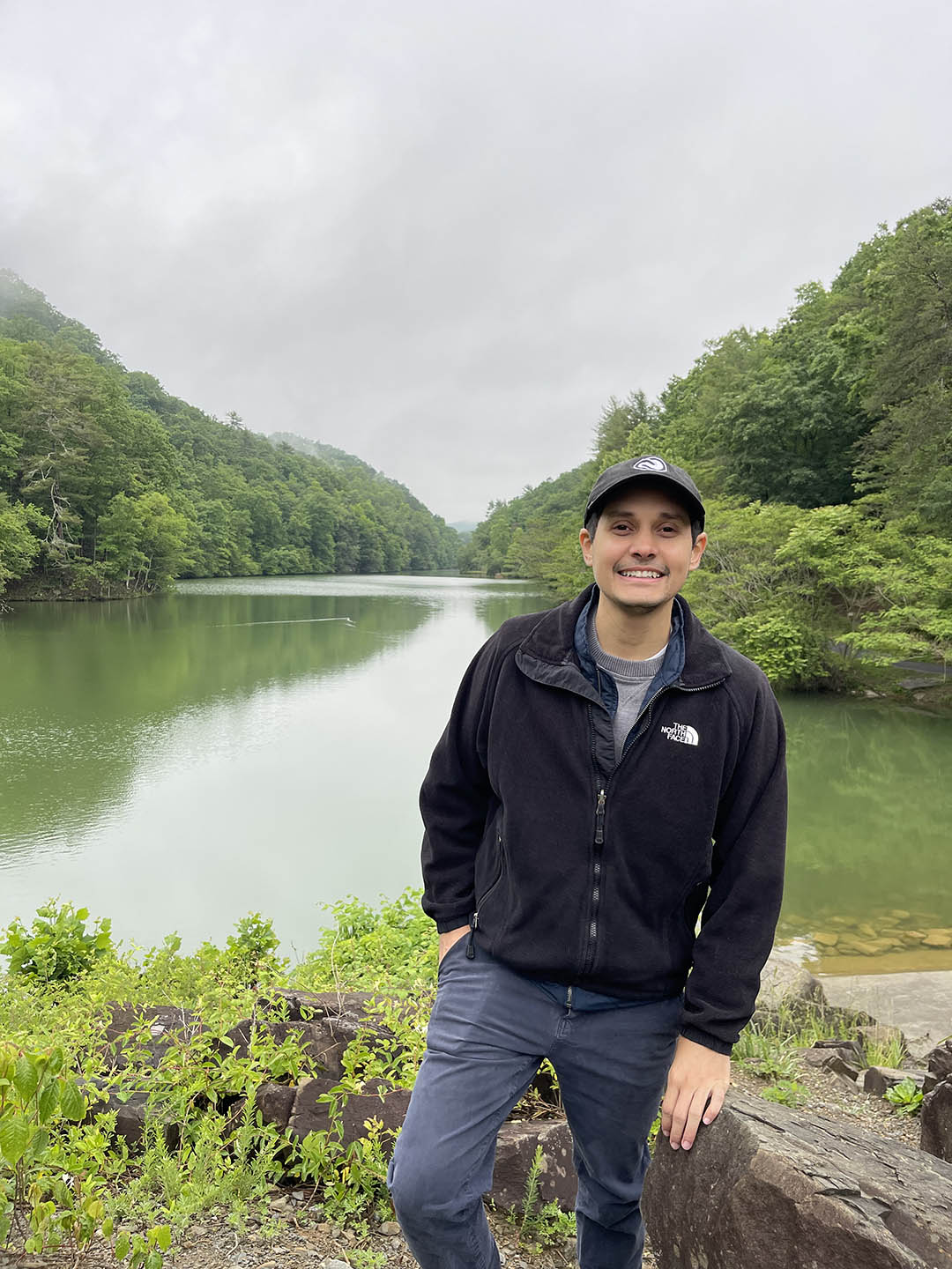 A photo of Michael Caraballo standing in front of a river with mountains and mist in the background.