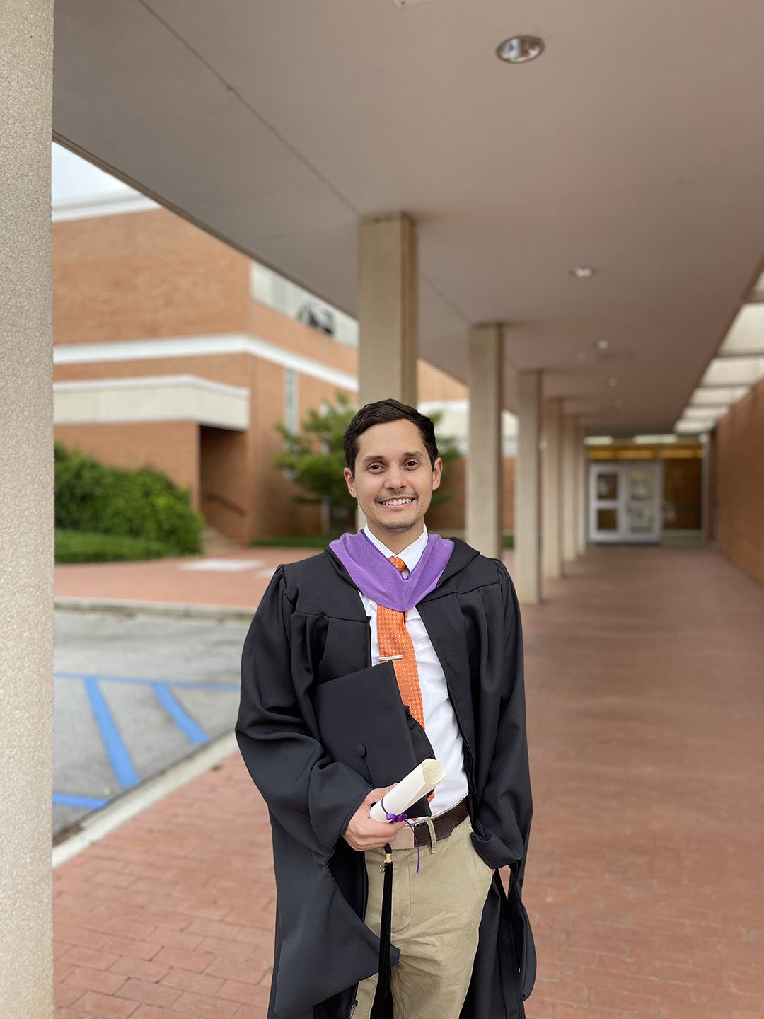 A photo of Michael Caraballo in his graduation cap and gown holding his diploma and standing in front of a brick building