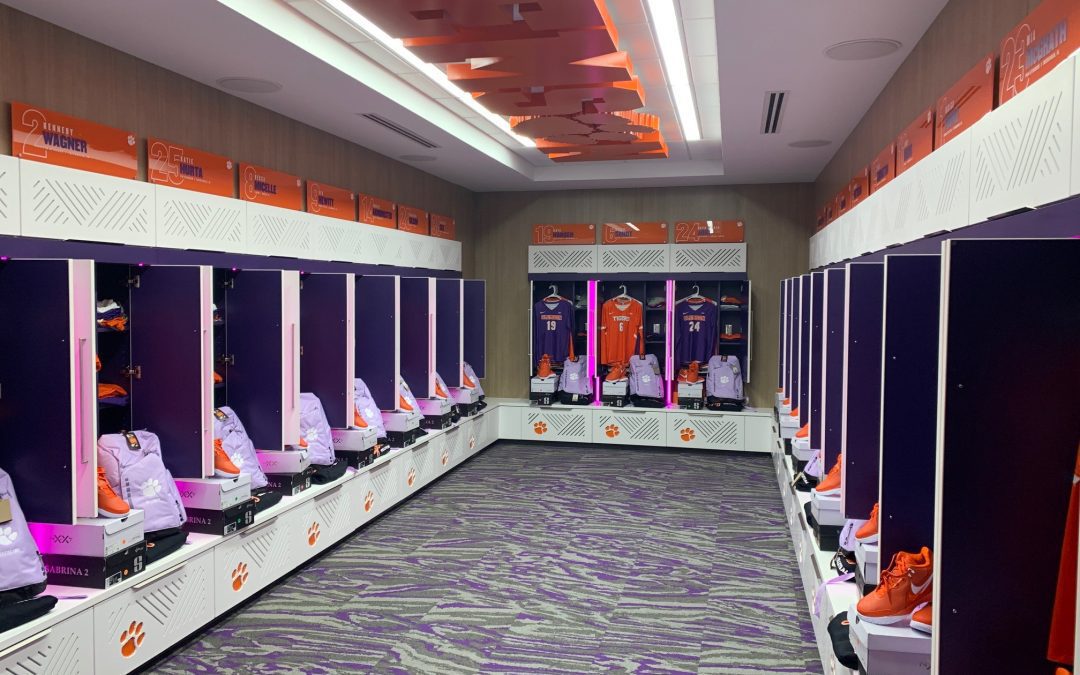 A photo of the new Clemson Volleyball Locker Room. Team jerseys and shoes are stacked inside white and purple lockers. Clemson is spelled out in orange block lettering on the ceiling above.