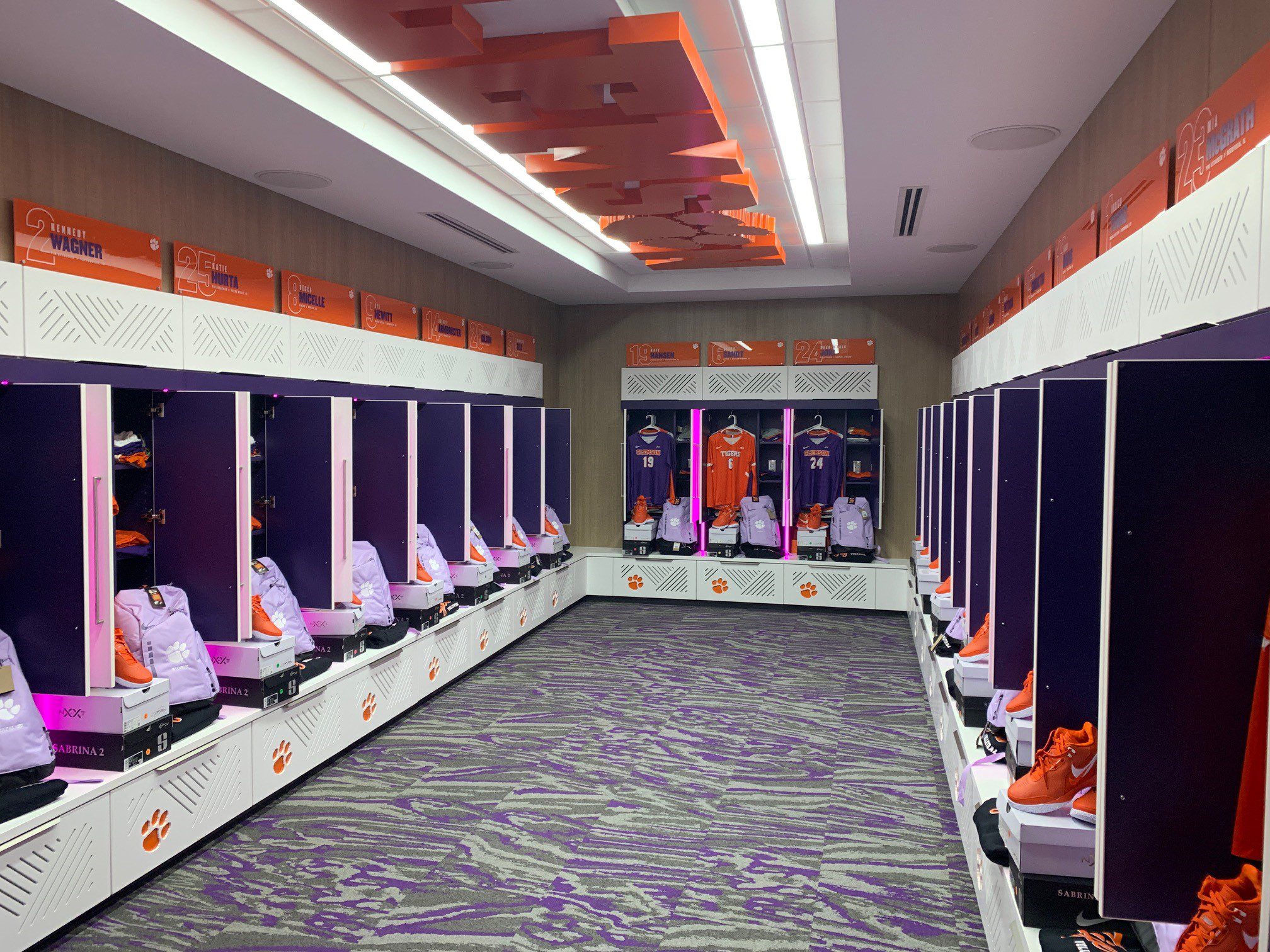 A photo of the new Clemson Volleyball Locker Room. Team jerseys and shoes are stacked inside white and purple lockers. Clemson is spelled out in orange block lettering on the ceiling above.