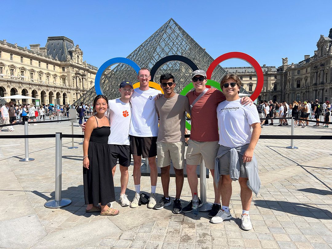 A photo of six young people standing in front of the Olympic rings outside of IM Pei's glass triangle at the Louvre in Paris.