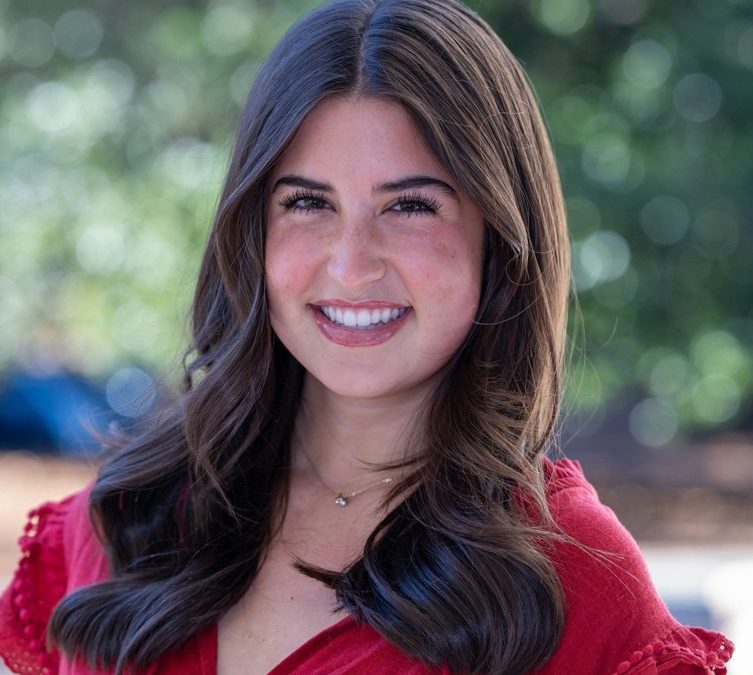 A photo of Kate Hughes, a young woman with long dark hair wearing a red dress and smiling in front of a group of trees.